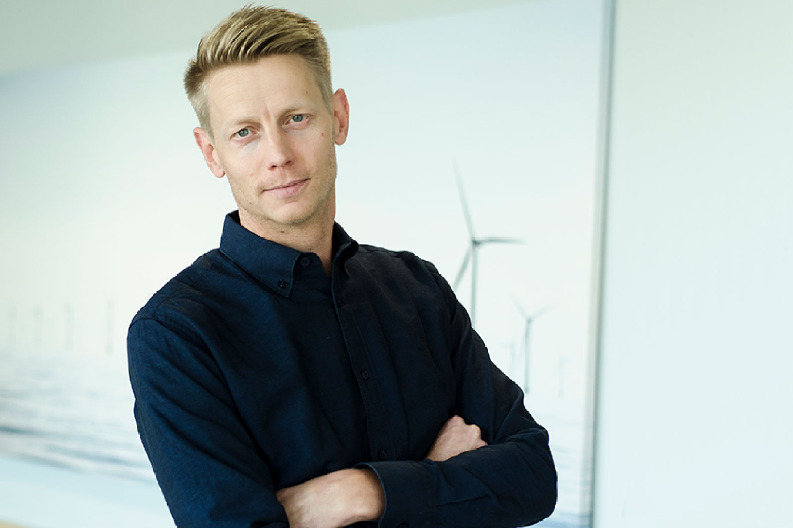 A person stands confidently with arms crossed, wearing a dark shirt, against a blurred backdrop featuring wind turbines.