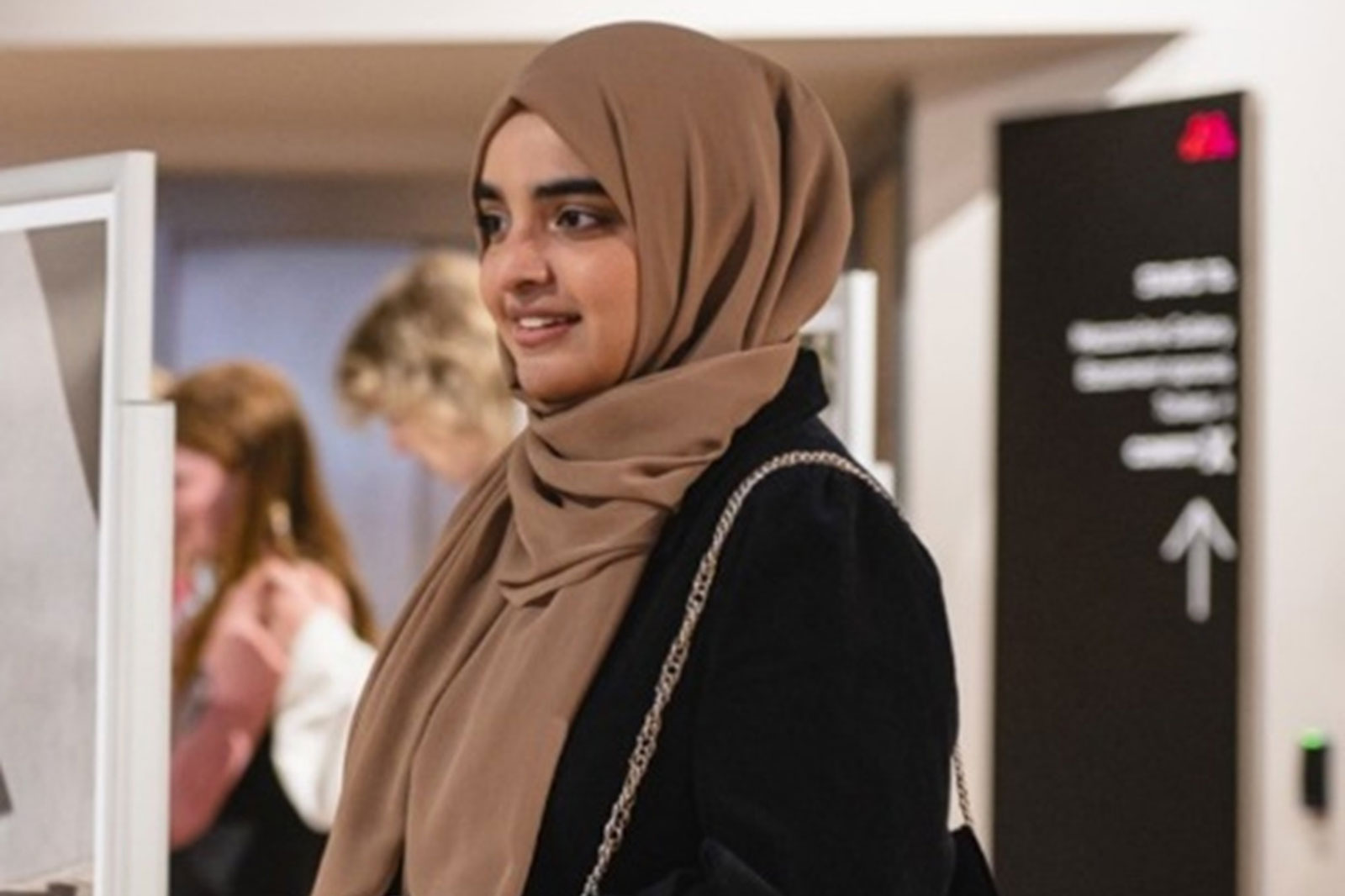 A woman wearing a brown hijab and a black jacket stands near a sign in a busy indoor space.
