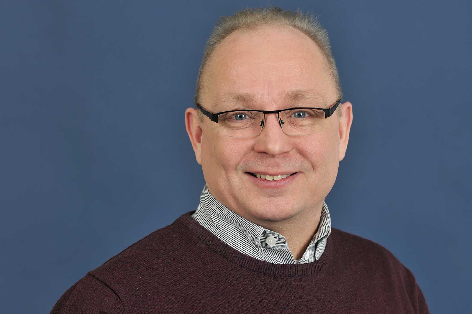 Eine Person mit einem dunklen Pulli und einem gestreiften Hemd steht vor einem blauen Hintergrund.