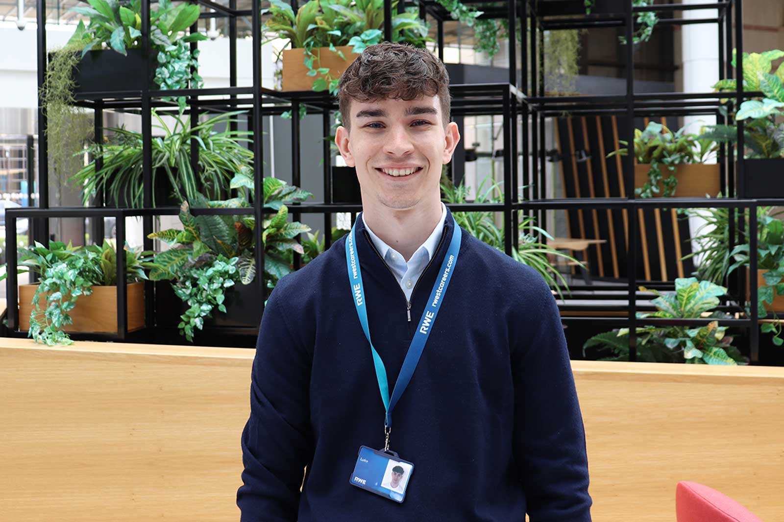 A person in a navy sweater stands in front of a green plant backdrop with wooden elements and a visitor badge.