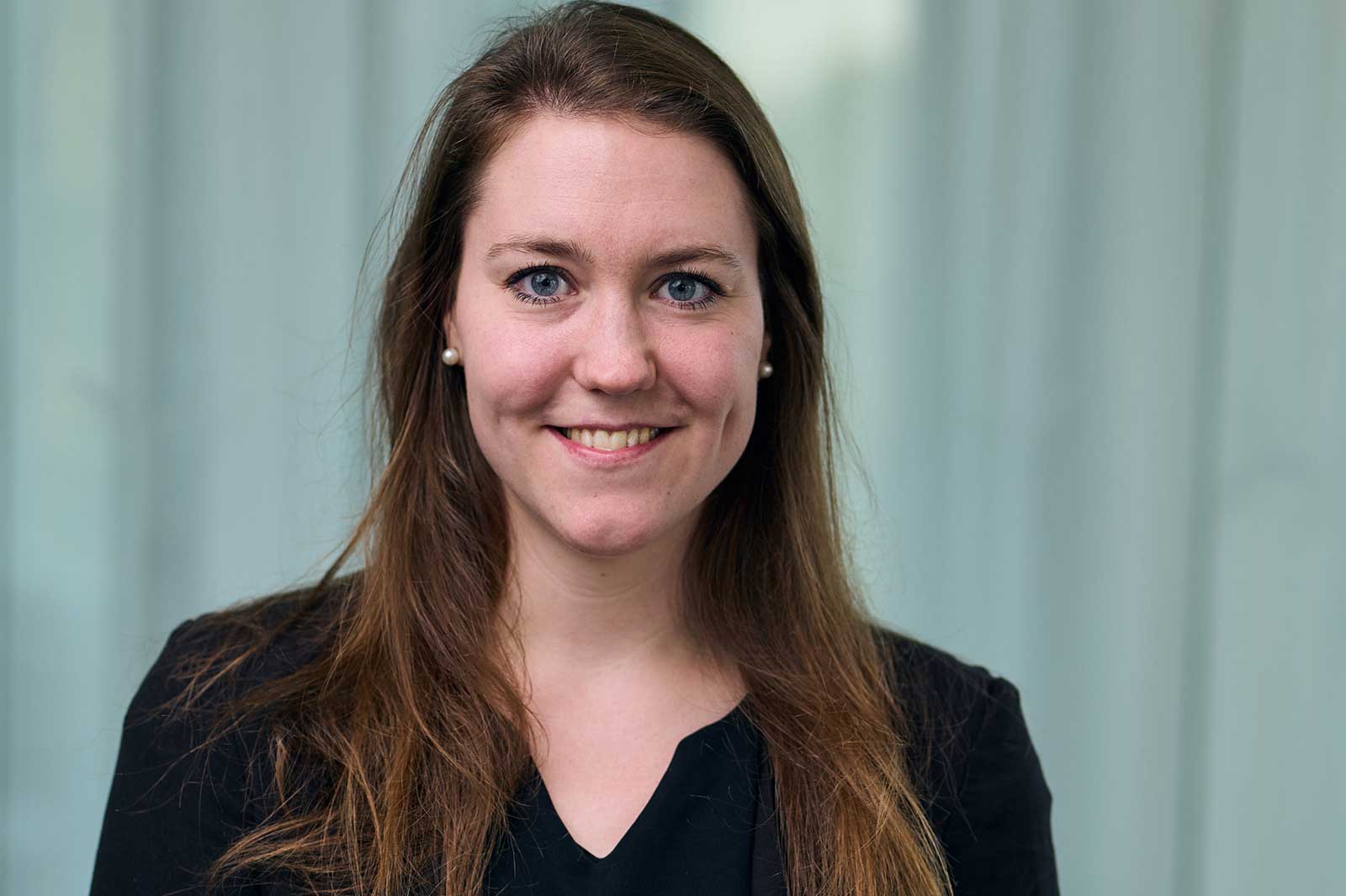 A woman with long brown hair, wearing a black top and pearl earrings, stands against a light green background.