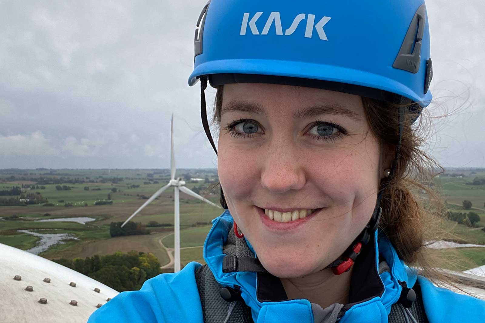 A person wearing a blue KASK helmet stands atop a wind turbine, overlooking green fields and a wind farm.