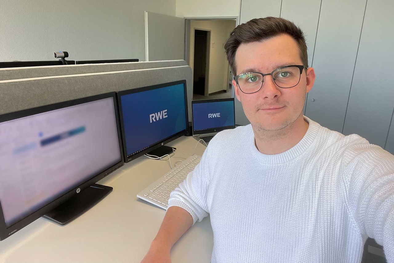 Eine Person in einem weißen Pullover posiert in einem Büro mit drei Computerbildschirmen, die das RWE-Logo zeigen.