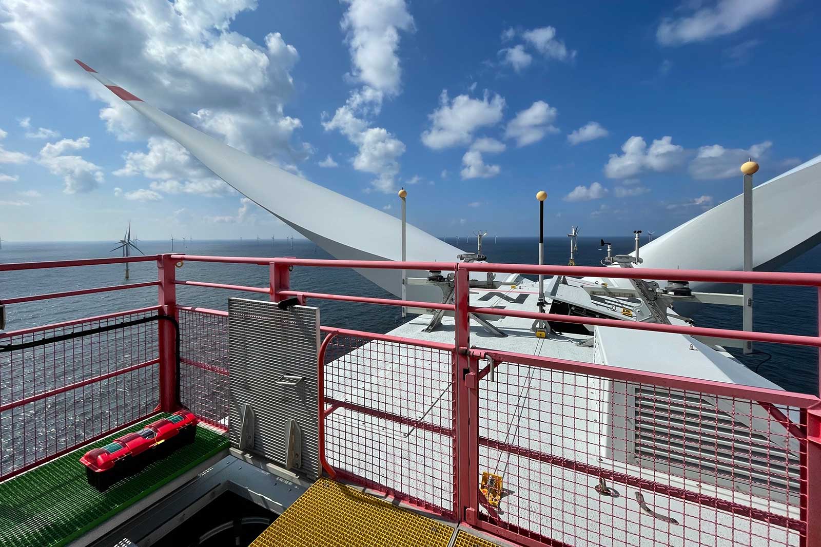 View from the top of a wind turbine over open water, with additional turbines in the background under a blue sky.