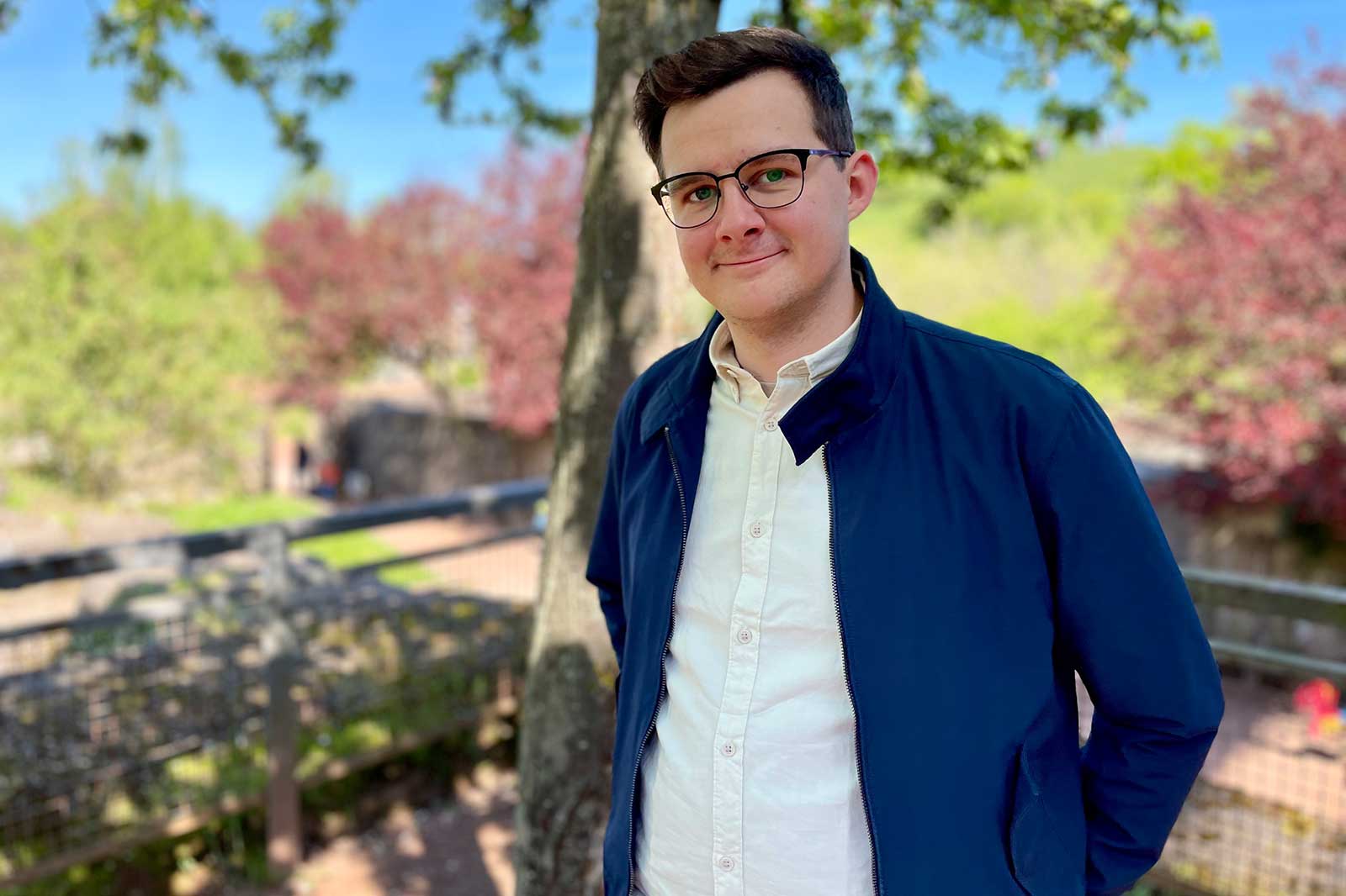 A person in a blue jacket stands outdoors before a green landscape with blossoming trees.