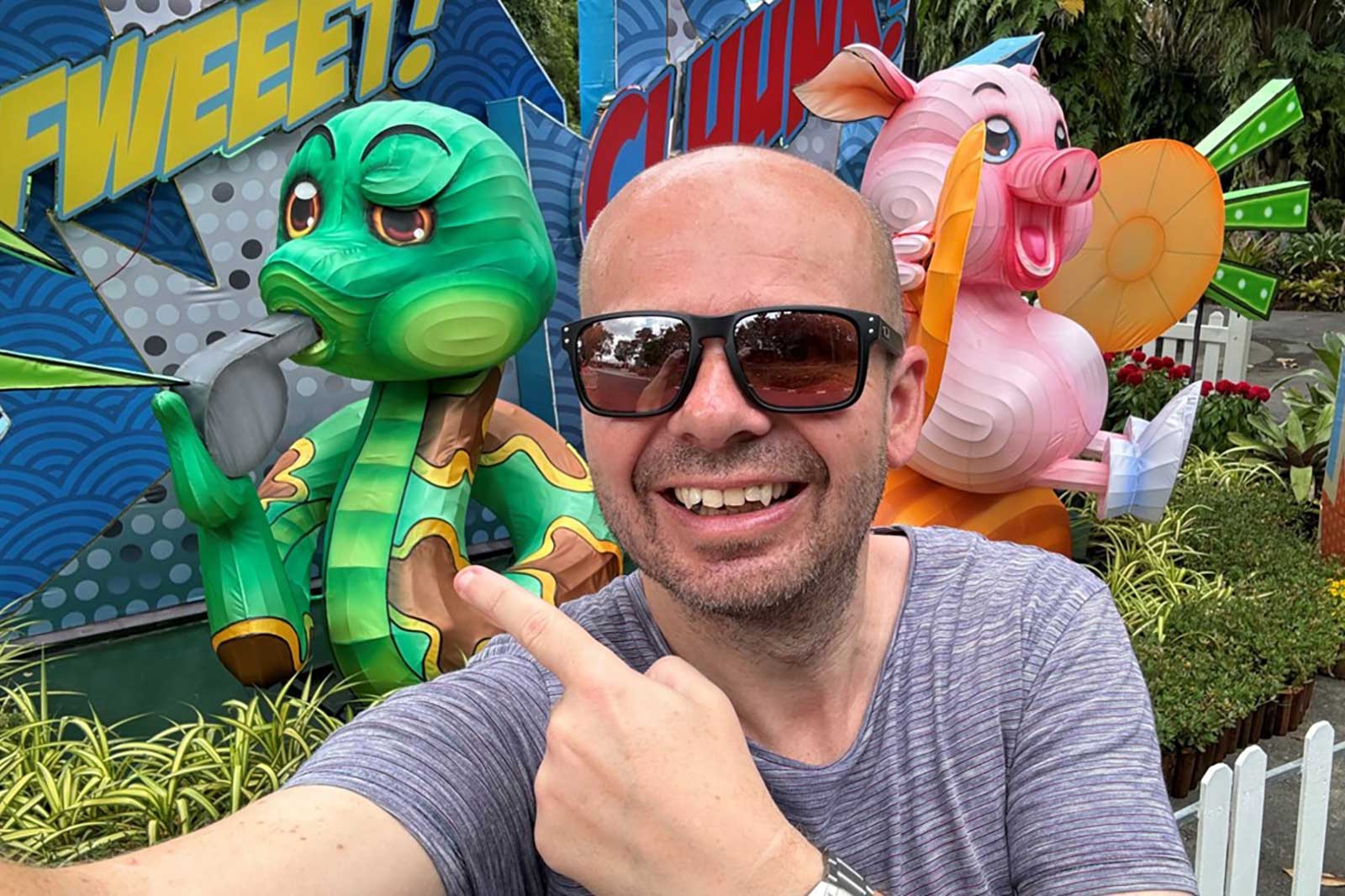 Ein grüner, cartoonartiger Drache, der Pfeife raucht, steht neben einem fröhlichen rosa Schwein mit einem Windrad.