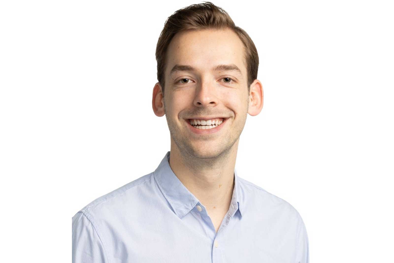 A man is wearing a light blue shirt and standing against a white background.