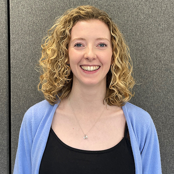 A smiling person with shoulder-length curly blonde hair wearing a light blue cardigan over a black top, standing before a gray fabric panel.