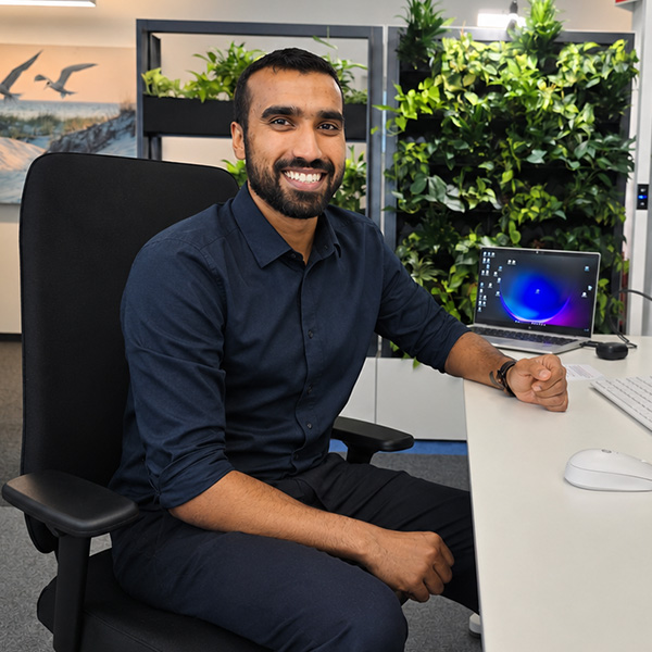 A smiling man with dark hair and beard sits at a white desk in a navy shirt, with a laptop and vertical green plant wall behind him.