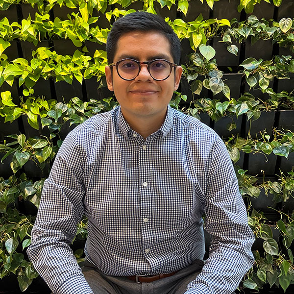 A smiling man sits facing the camera wearing round dark glasses and a black-and-white checked button-up shirt against a vertical green wall of plants.