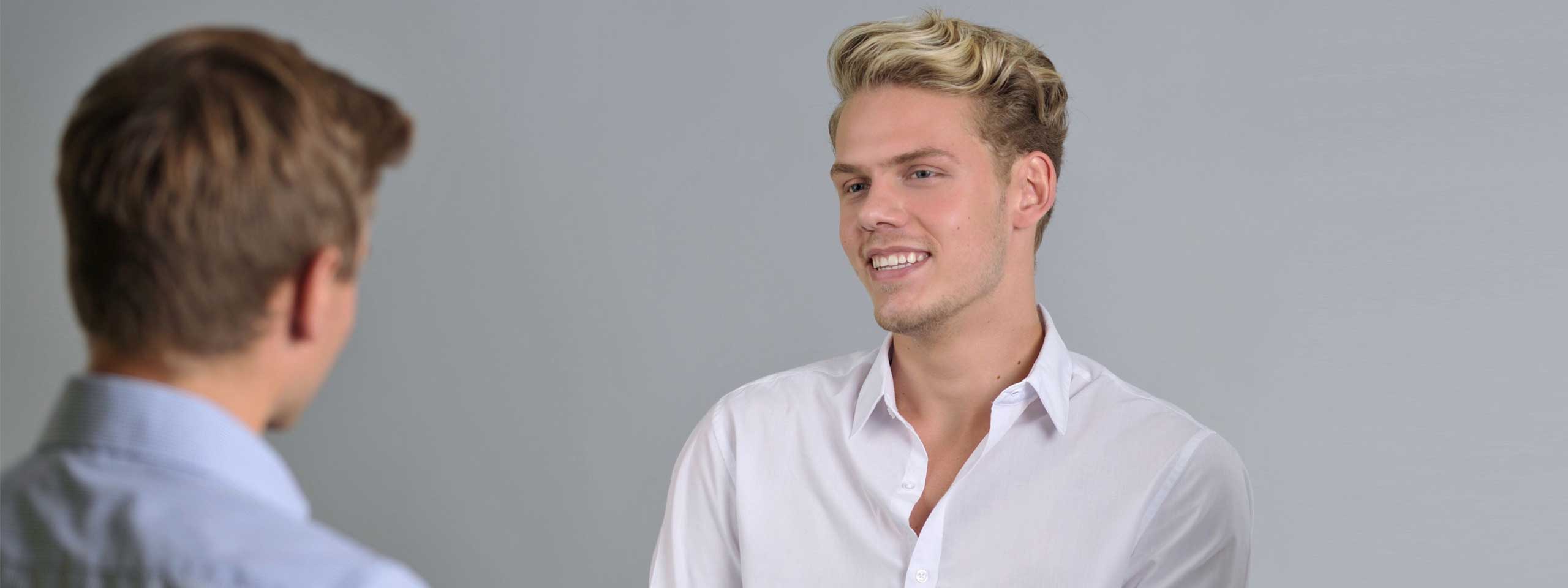 Zwei Personen führen ein Gespräch, einer trägt ein helles Hemd, vor einem glatten grauen Hintergrund.