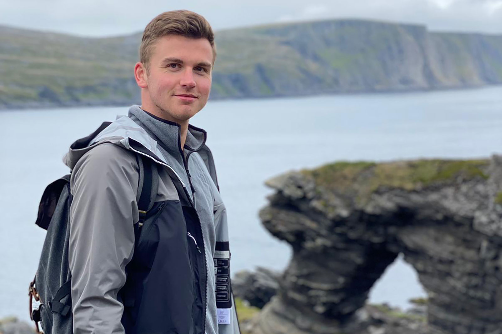 A man stands by the water's edge, with a striking rock formation in the background and green hills in the distance.