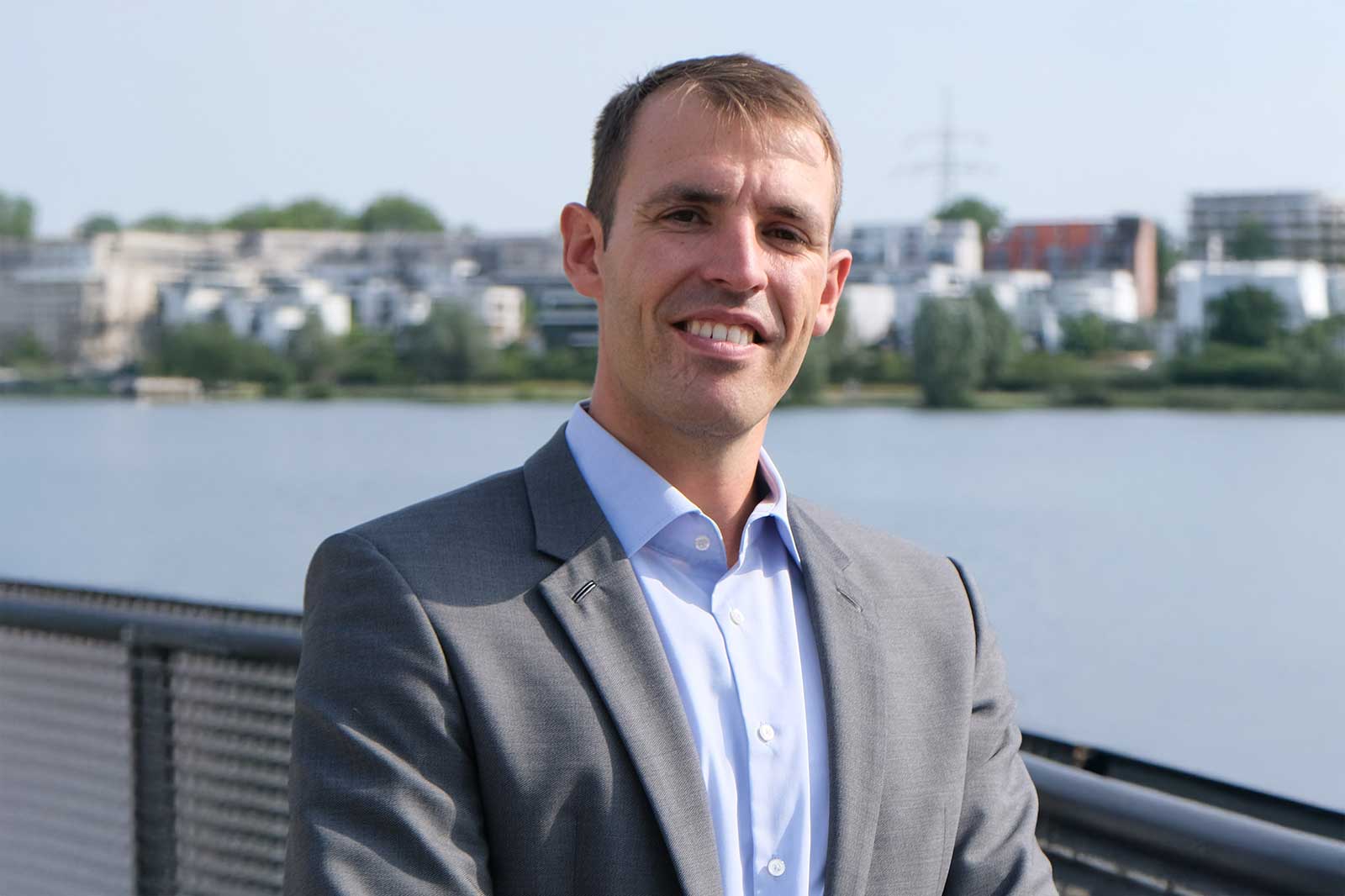 A man wearing a grey suit stands near a calm water body with modern buildings in the background on a sunny day.