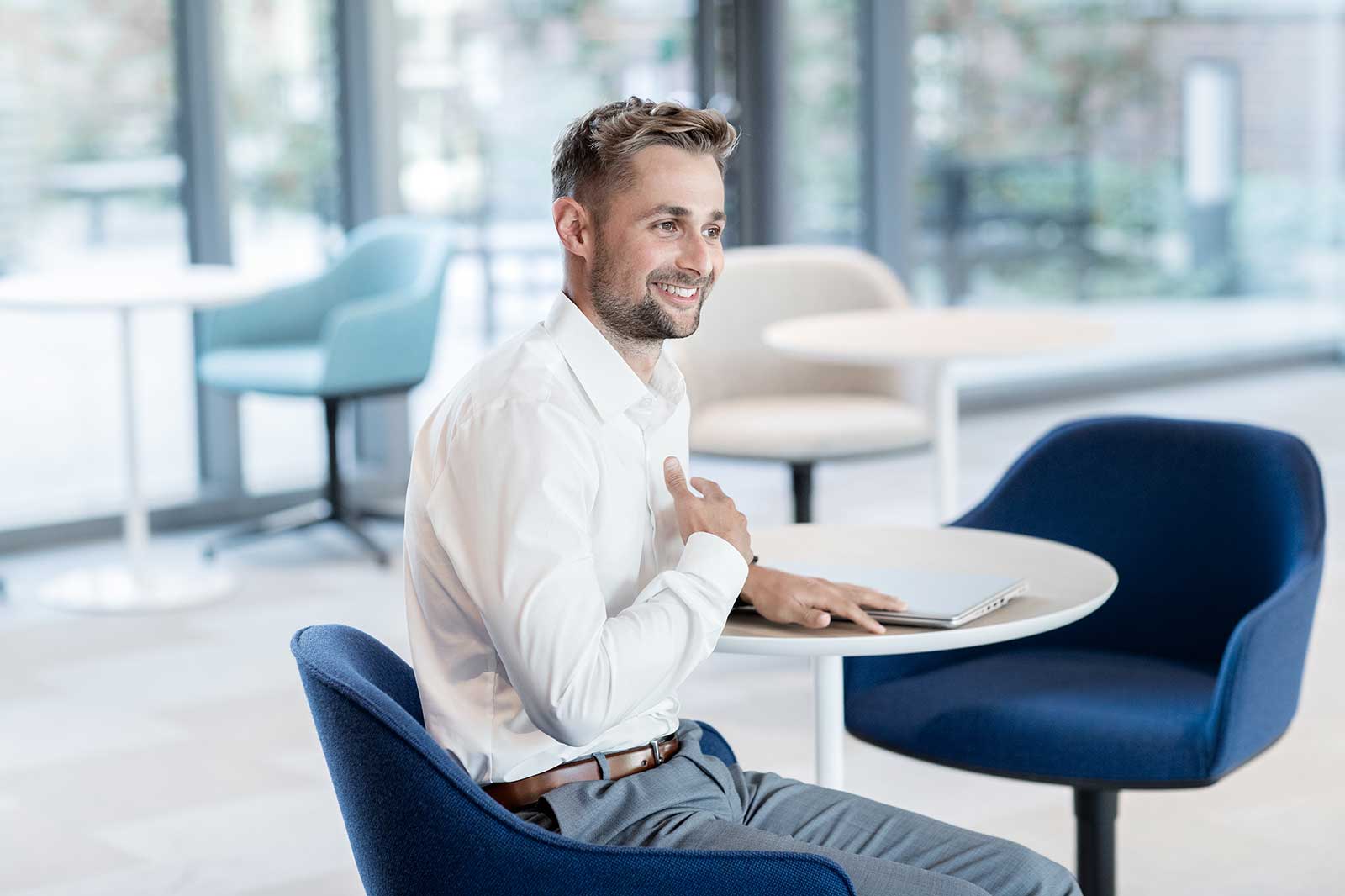 A seated man in a white shirt gestures while sitting at a round table in a modern café setting with blue chairs.
