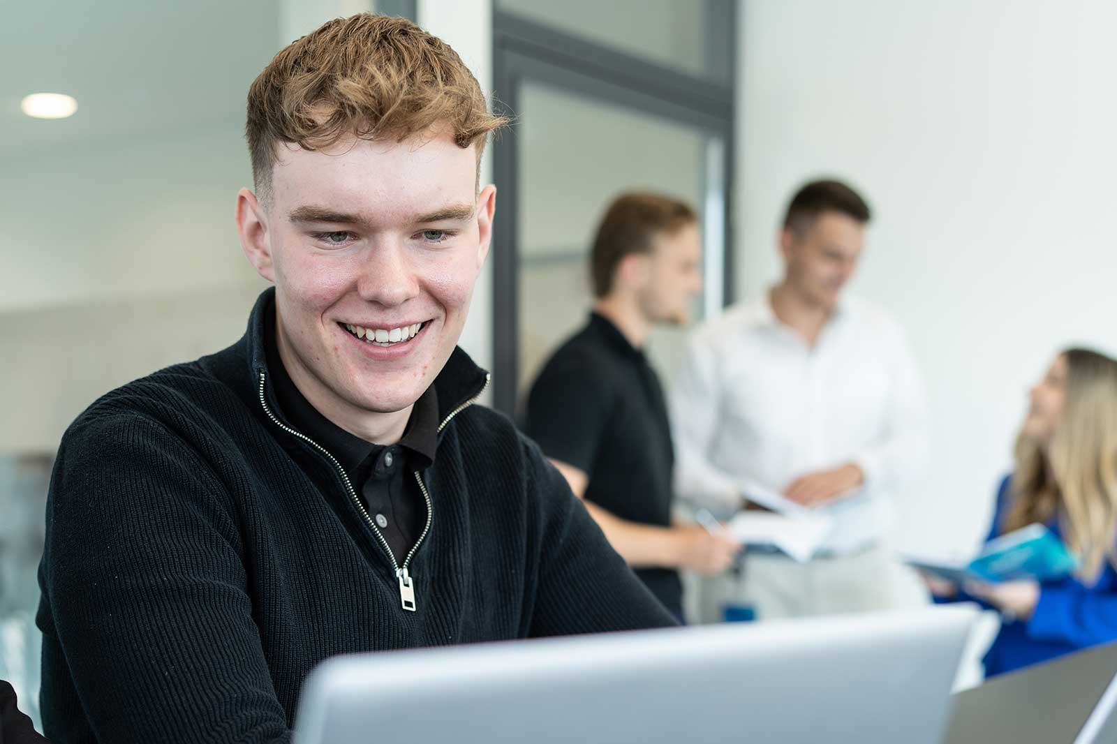 Ein junger Mann in einem schwarzen Pullover arbeitet an einem Laptop, während eine Gruppe von Kollegen im Hintergrund diskutiert.
