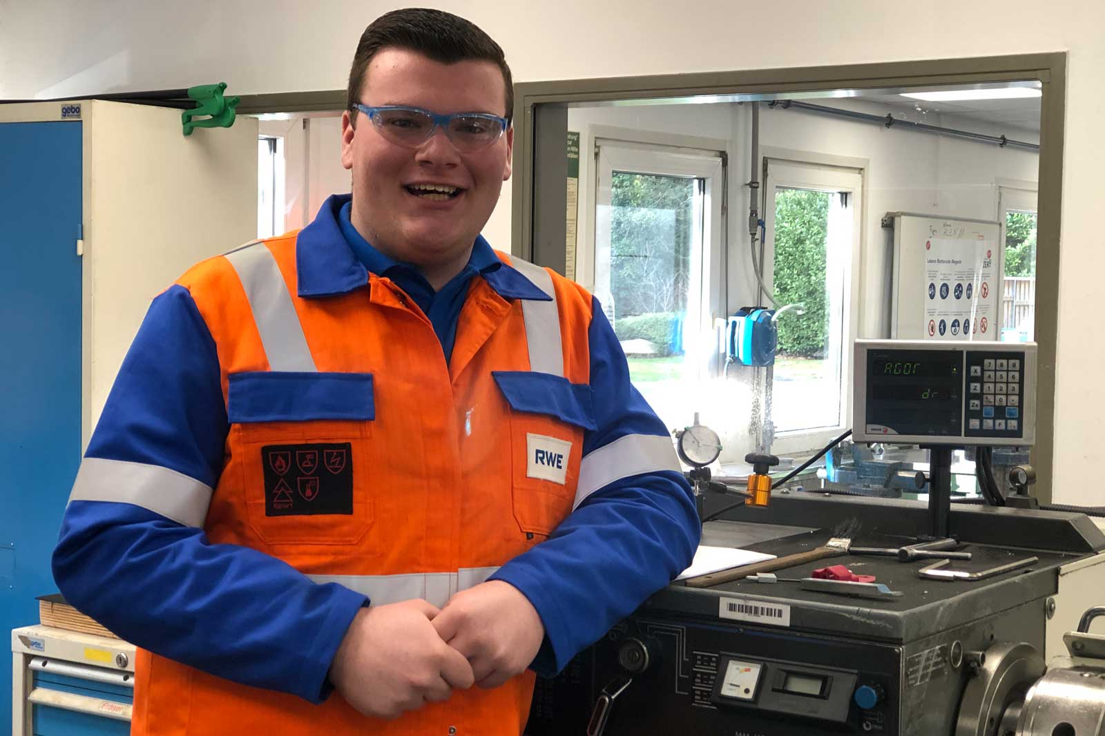 Ein Arbeiter in einer orange-blauen Uniform steht in einer Werkstatt, mit Ausrüstung und einem Monitor im Hintergrund.