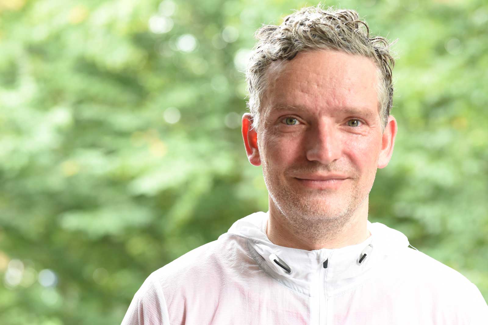 Ein Mann steht vor einem verschwommenen, grünen Hintergrund. Er trägt eine weiße Jacke mit Reißverschluss.