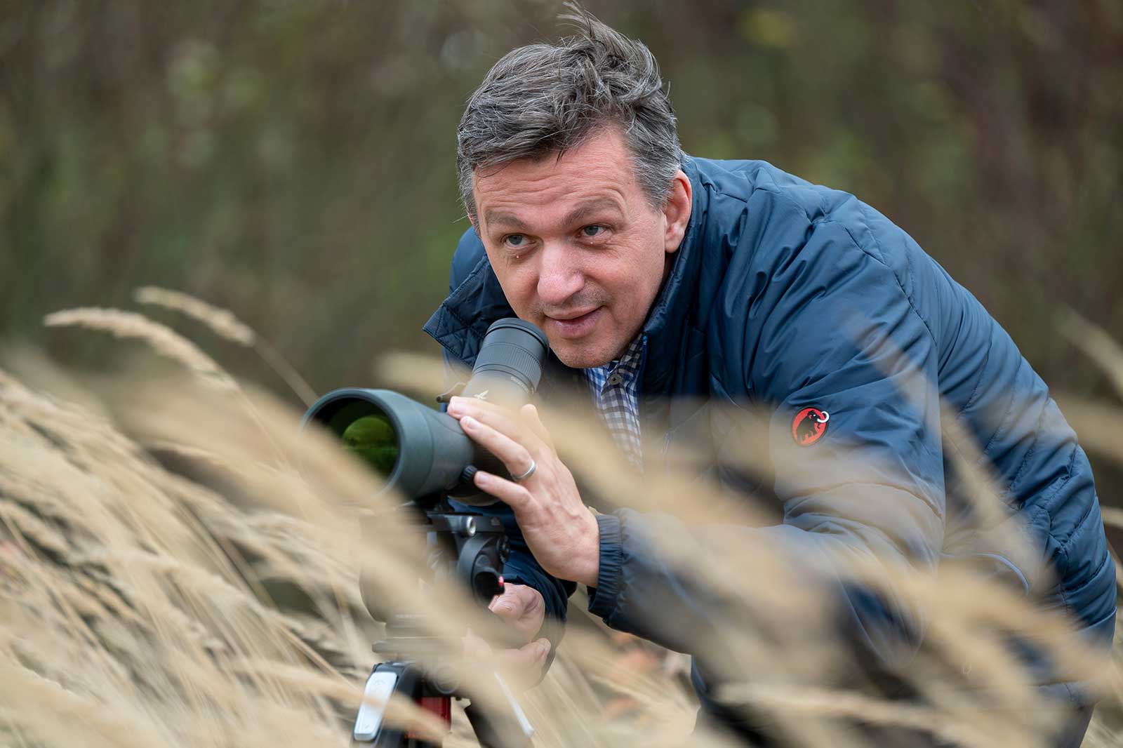 A man with a telescope is seated in tall grass, observing his surroundings attentively.