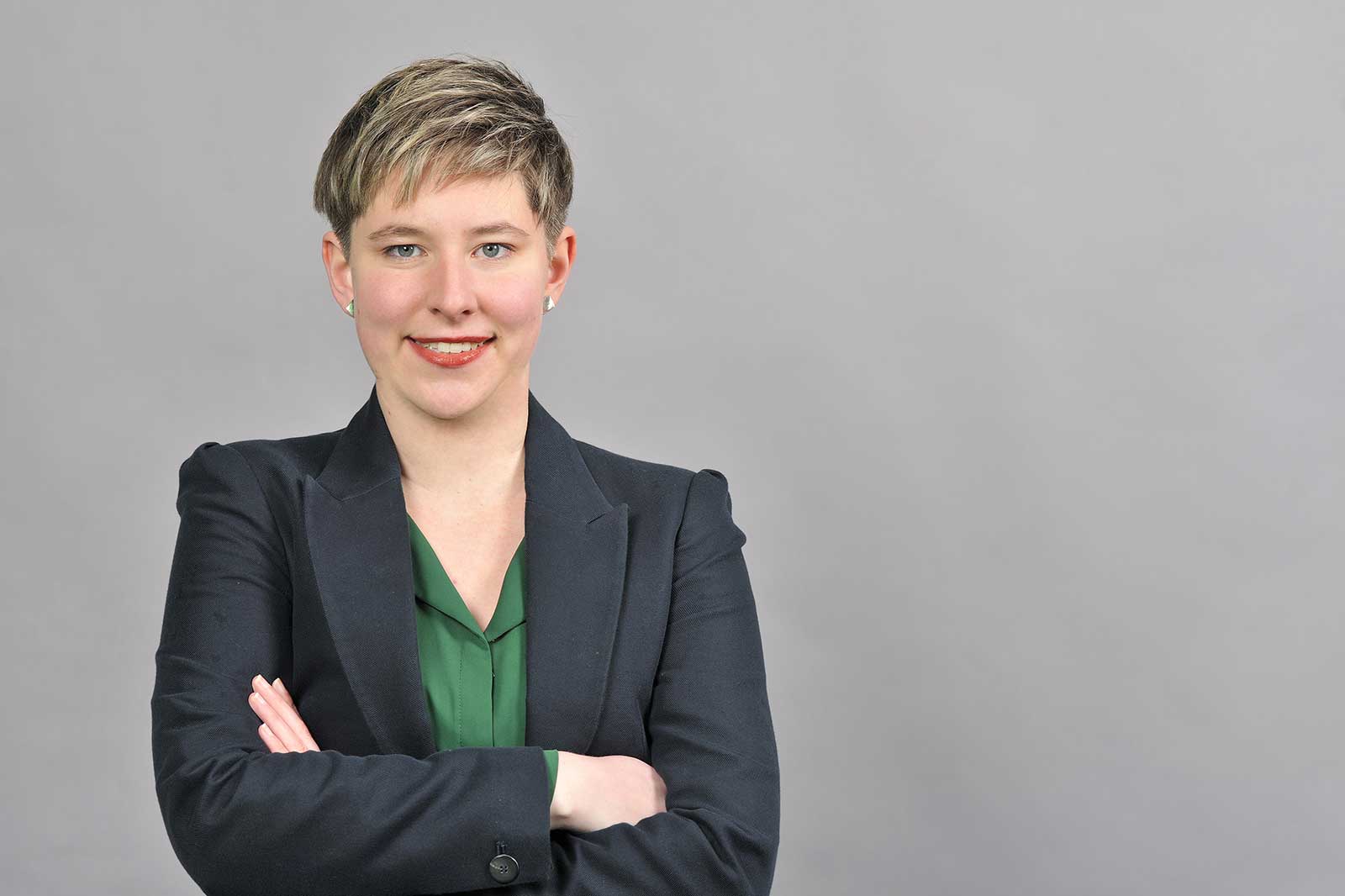 Eine Person in einem schwarzen Blazer mit verschränkten Armen vor einem grauen Hintergrund.