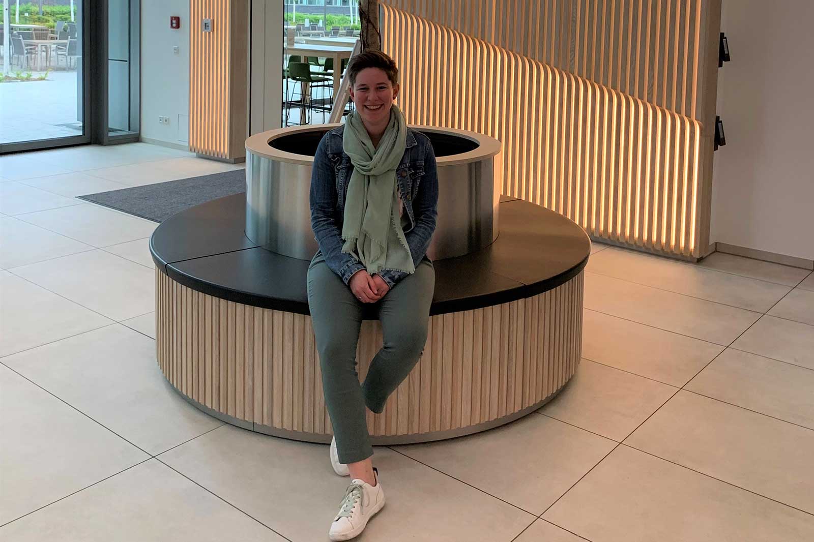 Eine Person sitzt auf einem runden Sitzbereich aus Holz und Schwarz in einem modernen Raum mit großen Fenstern.