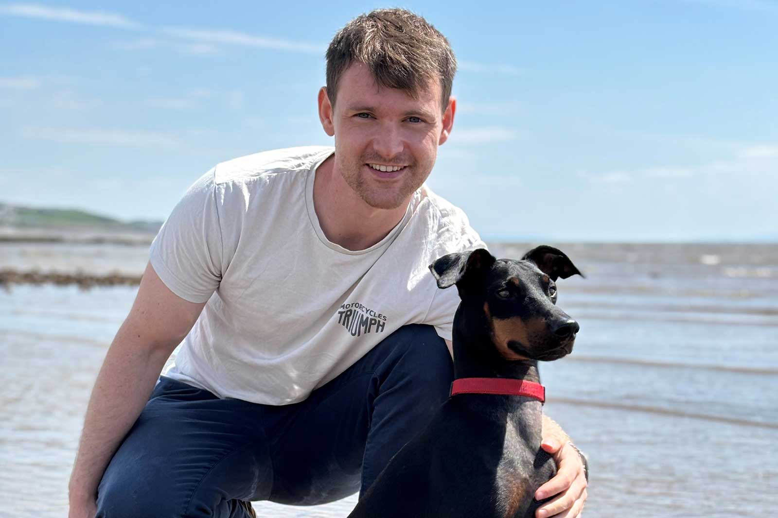 A man kneels on the beach next to a black dog. The sky is blue and the waves are gentle.