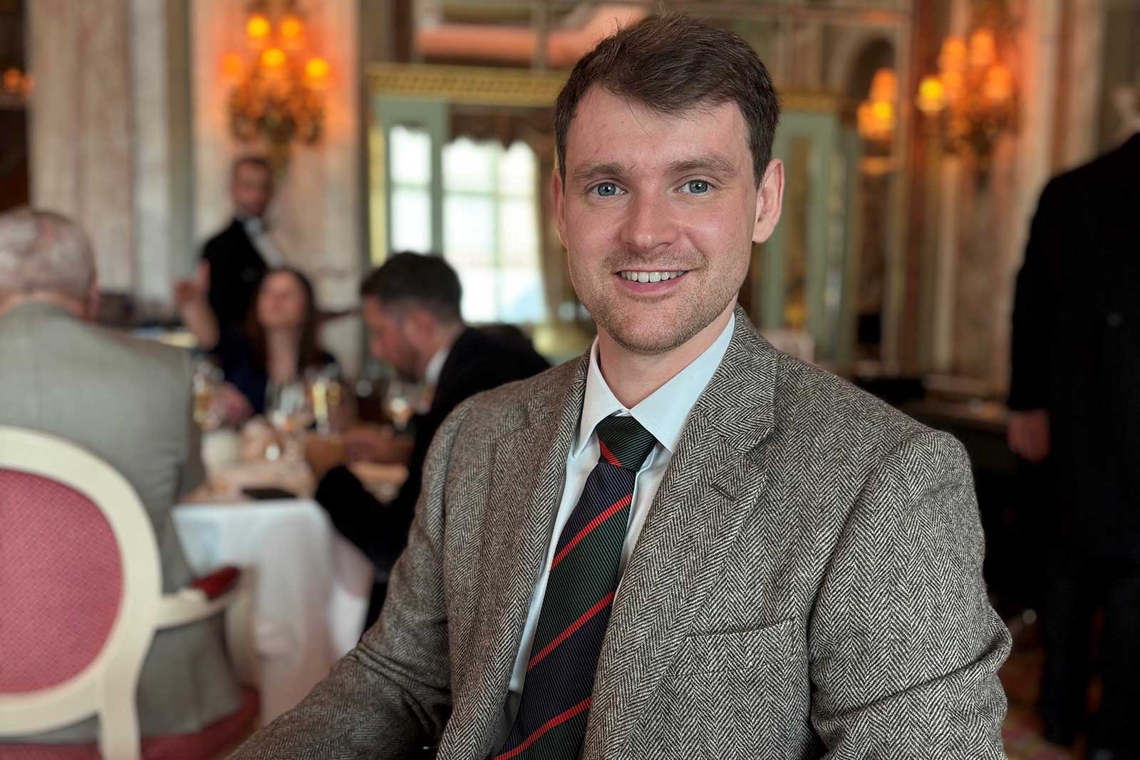 A man in an elegant restaurant, wearing a grey tweed suit with a striped tie, is seated at a table.