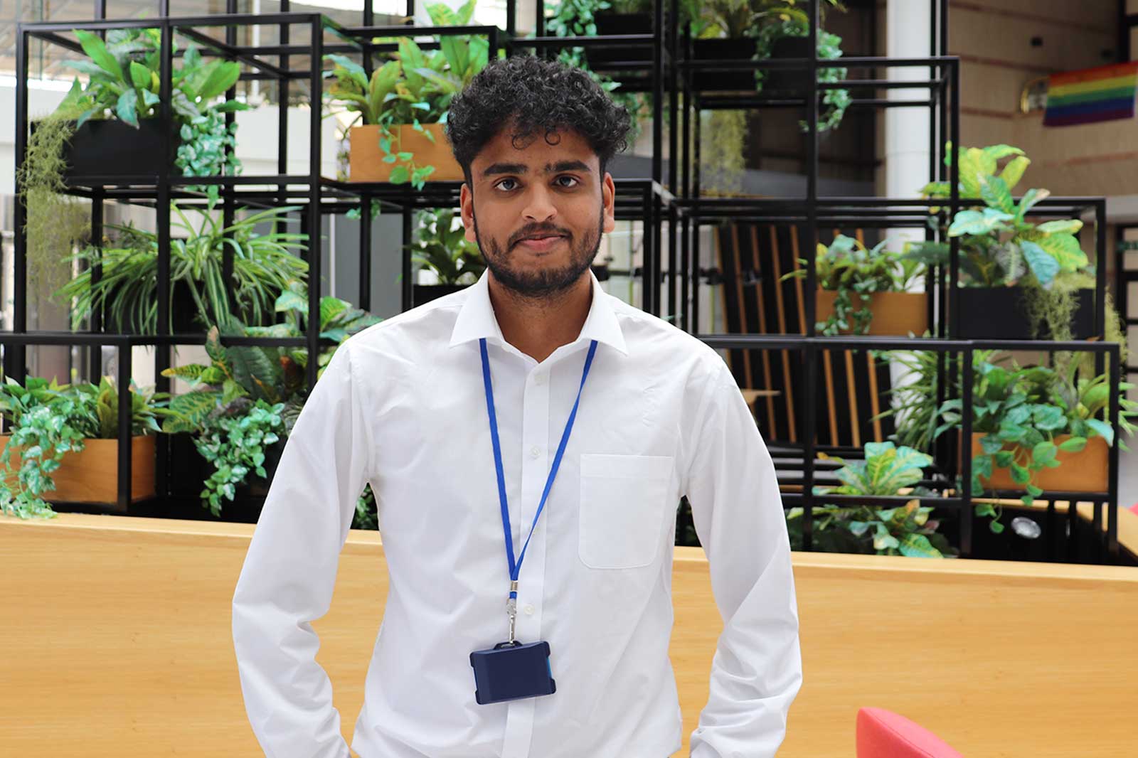 A person wearing a white shirt with a blue lanyard stands in front of a decorative plant display in a modern setting.