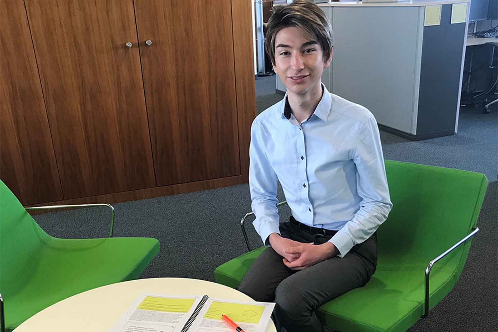 Eine sitzende Person in einem hellblauen Hemd und grauen Hosen. Grüne Stühle und ein runder Tisch mit Dokumenten und einem Stift.