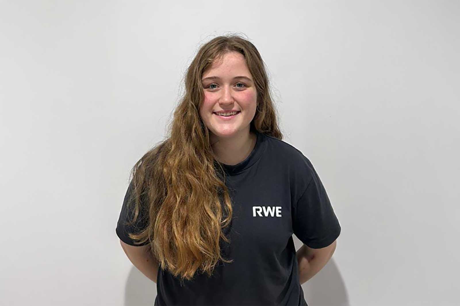 A person with long hair stands facing forward wearing a black t-shirt with 'RWE' printed in white.