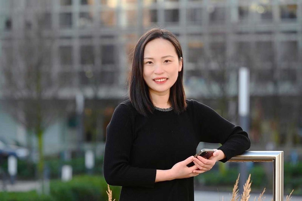 Eine Frau in einem schwarzen Pullover steht mit einem Smartphone in der Hand vor einem modernen städtischen Hintergrund.