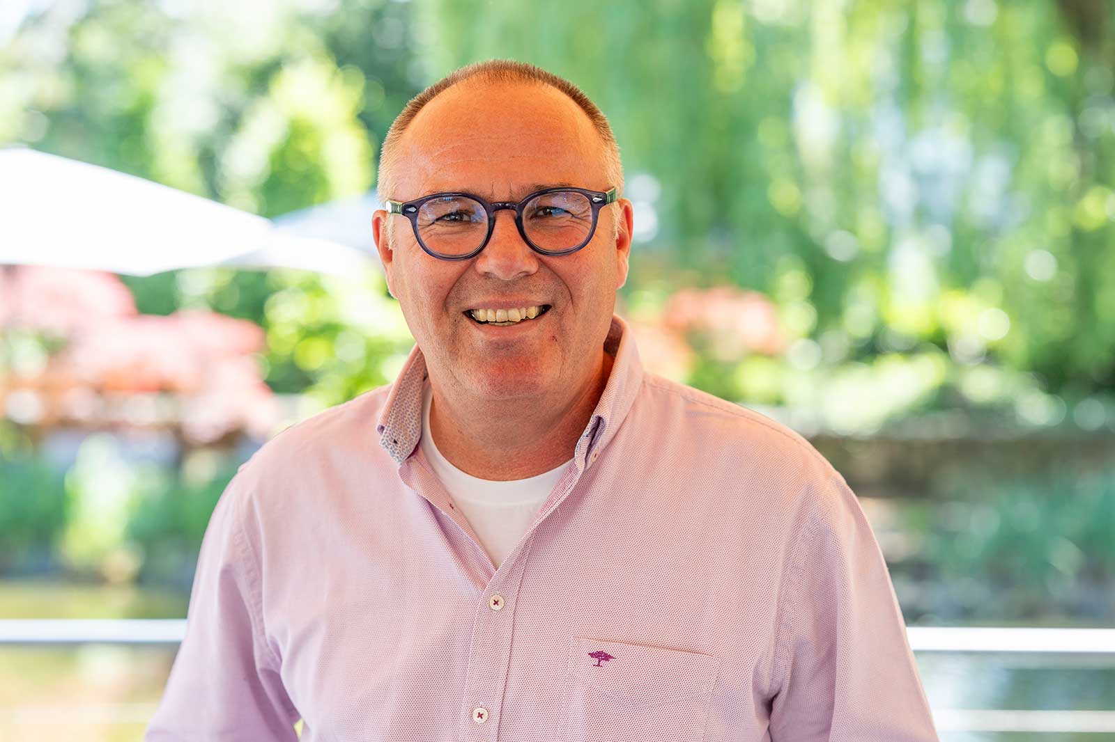 Ein Mann mit kurz geschnittenem Haar und sichtbaren Brillenbügeln trägt ein rosa Hemd über einem weißen T-Shirt.