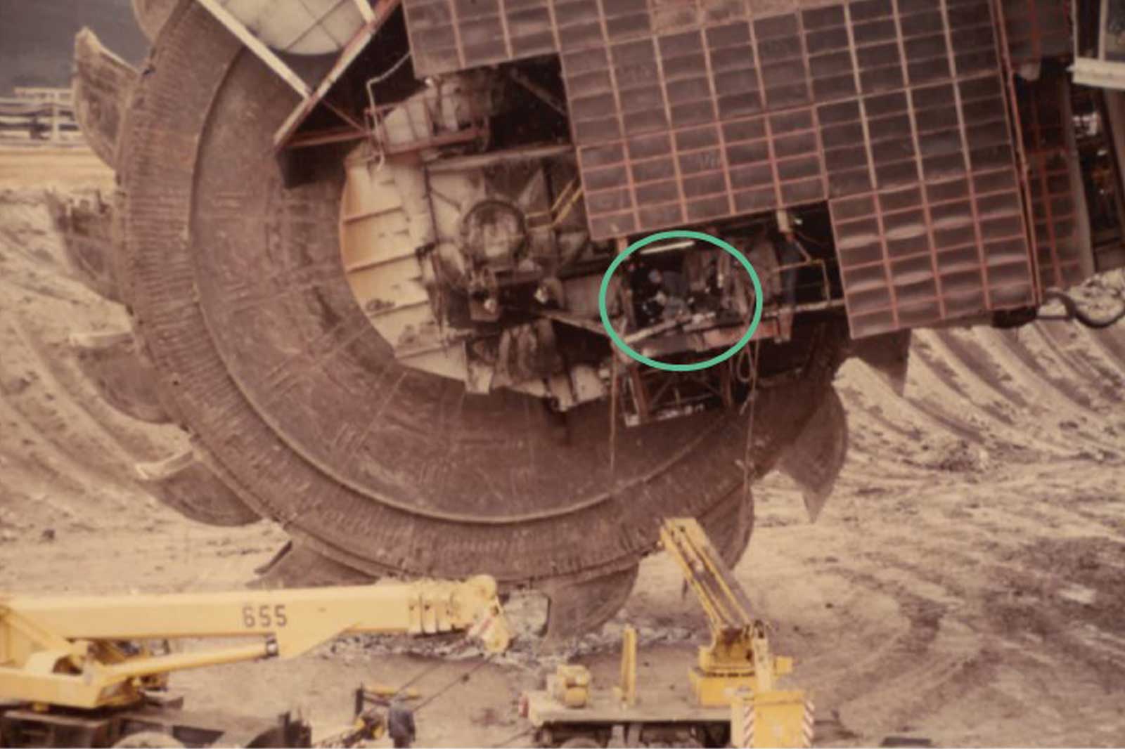 Ein großer Bagger mit einer riesigen rotierenden Trommel, die Arbeiter beim Reparieren von Maschinen an einem Bergbauort zeigt.