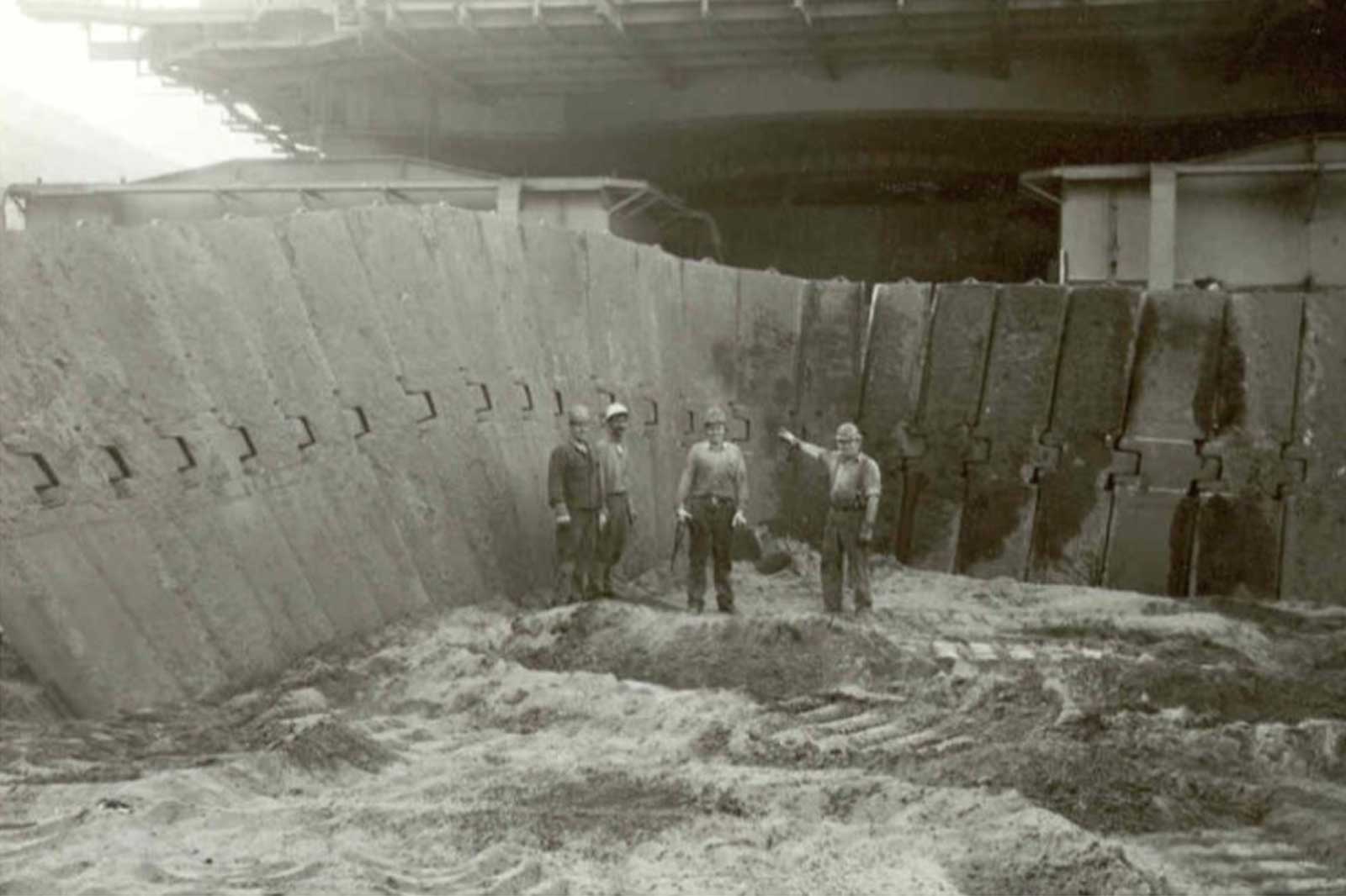 Arbeiter stehen an einer Baustelle mit einer großen Betonwand und sandigem Boden.