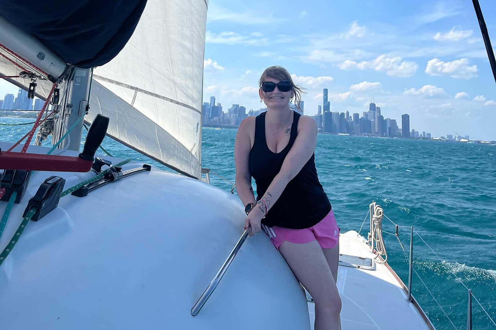 Eine Person steht auf einem Segelboot mit Blick auf die Stadt im Hintergrund. Der Himmel ist blau mit einigen Wolken.