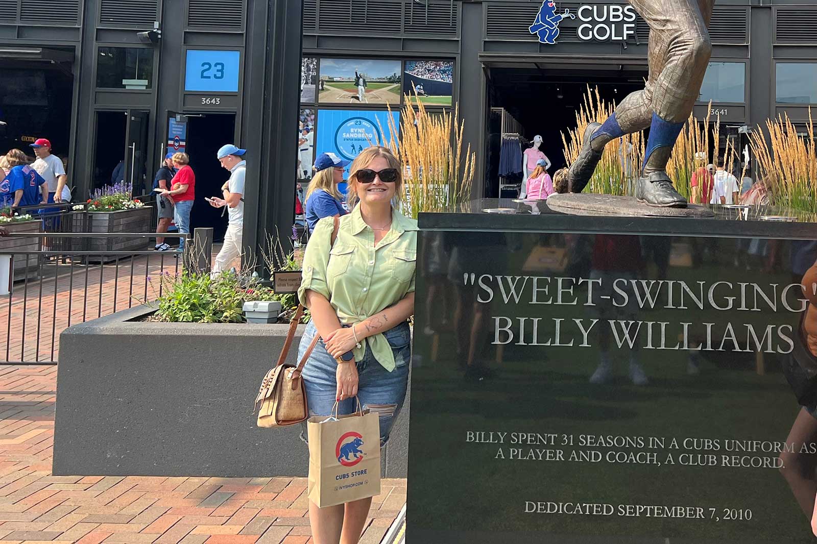 Eine Statue von Billy Williams steht vor dem Cubs Golf Store, umgeben von Menschen und Blumenbeeten.