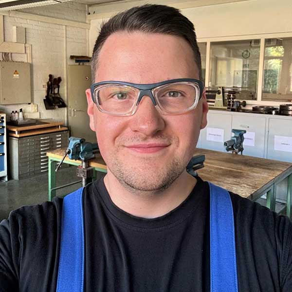 Eine Person in einem Werkstattumfeld trägt ein schwarzes T-Shirt und blaue Latzhosen. Im Hintergrund sind Werkbänke und Werkzeuge sichtbar.