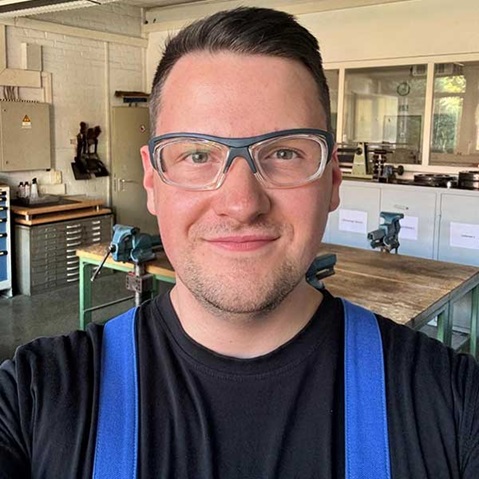 Eine Person in einem Werkstattumfeld trägt ein schwarzes T-Shirt und blaue Latzhosen. Im Hintergrund sind Werkbänke und Werkzeuge sichtbar.