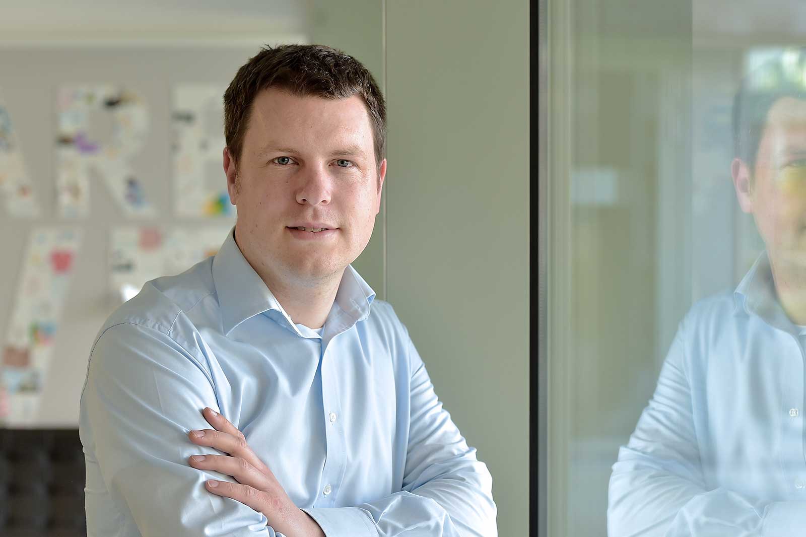 A man in a light blue shirt stands by a glass window, arms crossed, with colourful artwork in the background.