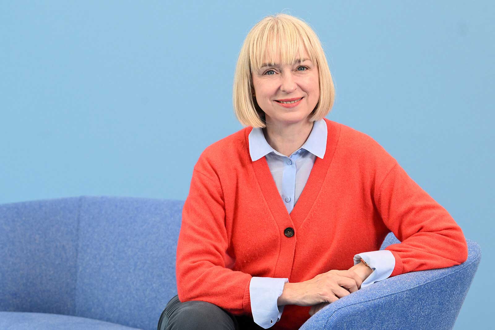 Eine Person sitzt auf einer blauen Couch, trägt einen roten Pullover und ein hellblaues Hemd vor einer blauen Wand.