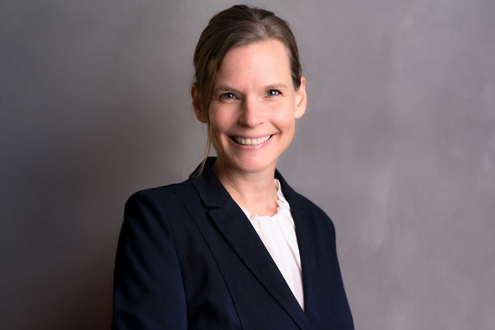Eine Frau in einem schwarzen Blazer mit einer weißen Bluse vor einem grauen Hintergrund.