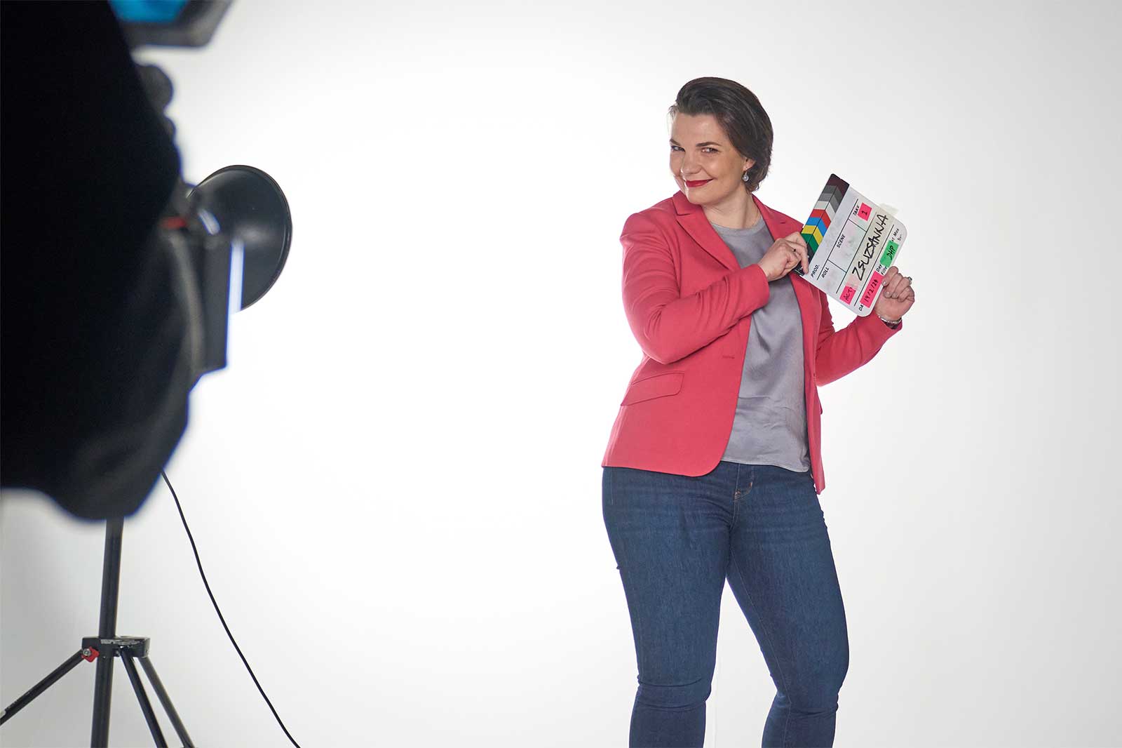 A person in a red blazer stands holding a clapperboard against a white background.