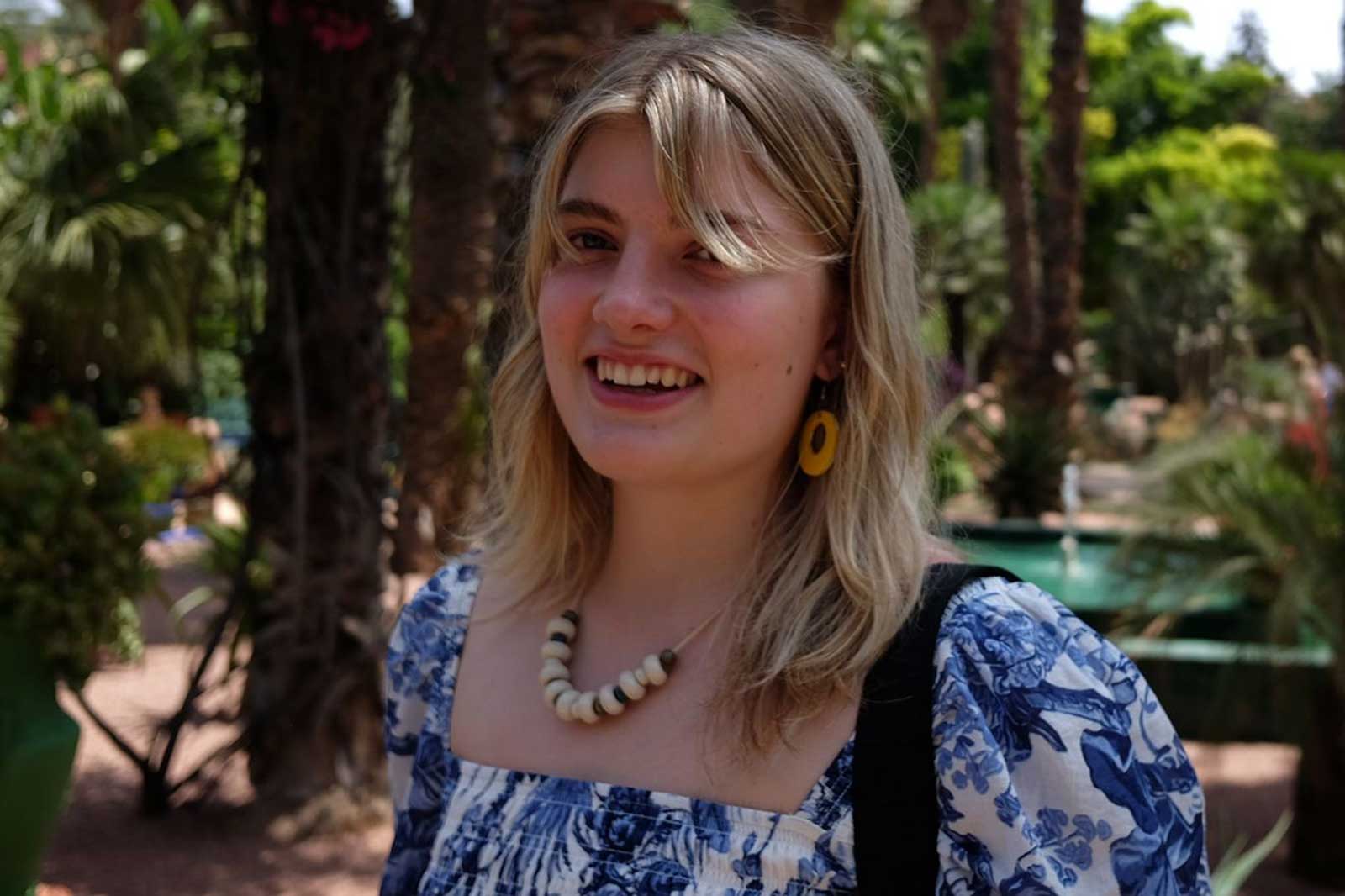 Eine Frau mit blonden Haaren trägt eine bunte Bluse und eine Halskette. Der Hintergrund zeigt Palmen und Pflanzen.