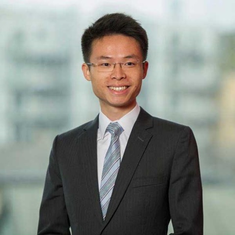 A man in a dark suit stands in front of a blurred urban background with a light backdrop.
