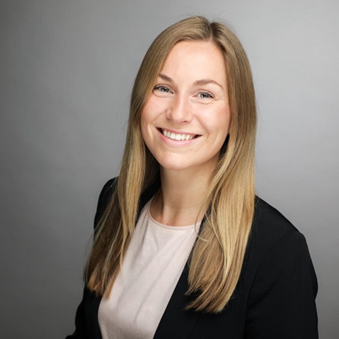 A person with long blonde hair wears a black blazer and a light top. The background is grey.