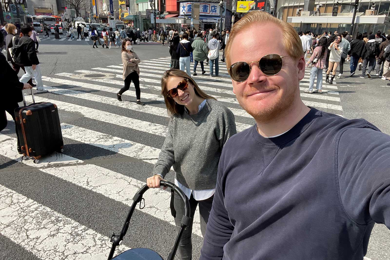 Two people stand at a crosswalk in a busy city. One person is pushing a stroller.