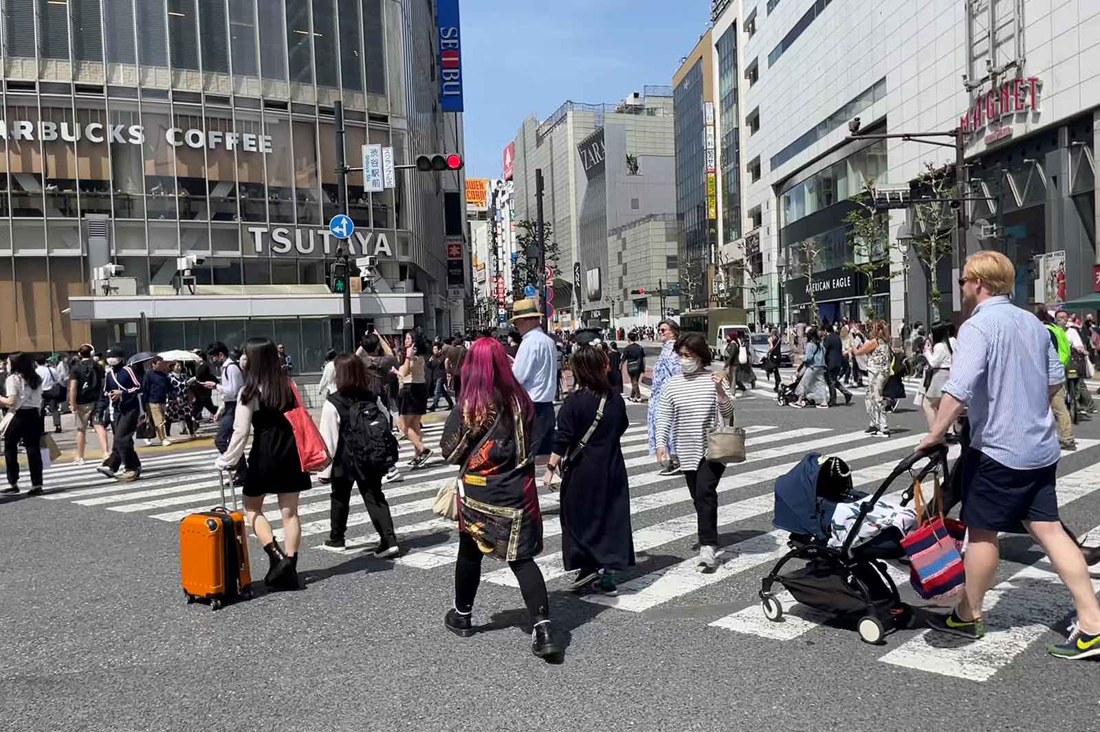 A busy urban crosswalk filled with pedestrians. Shops like Starbucks and Tsutaya are visible in the background.