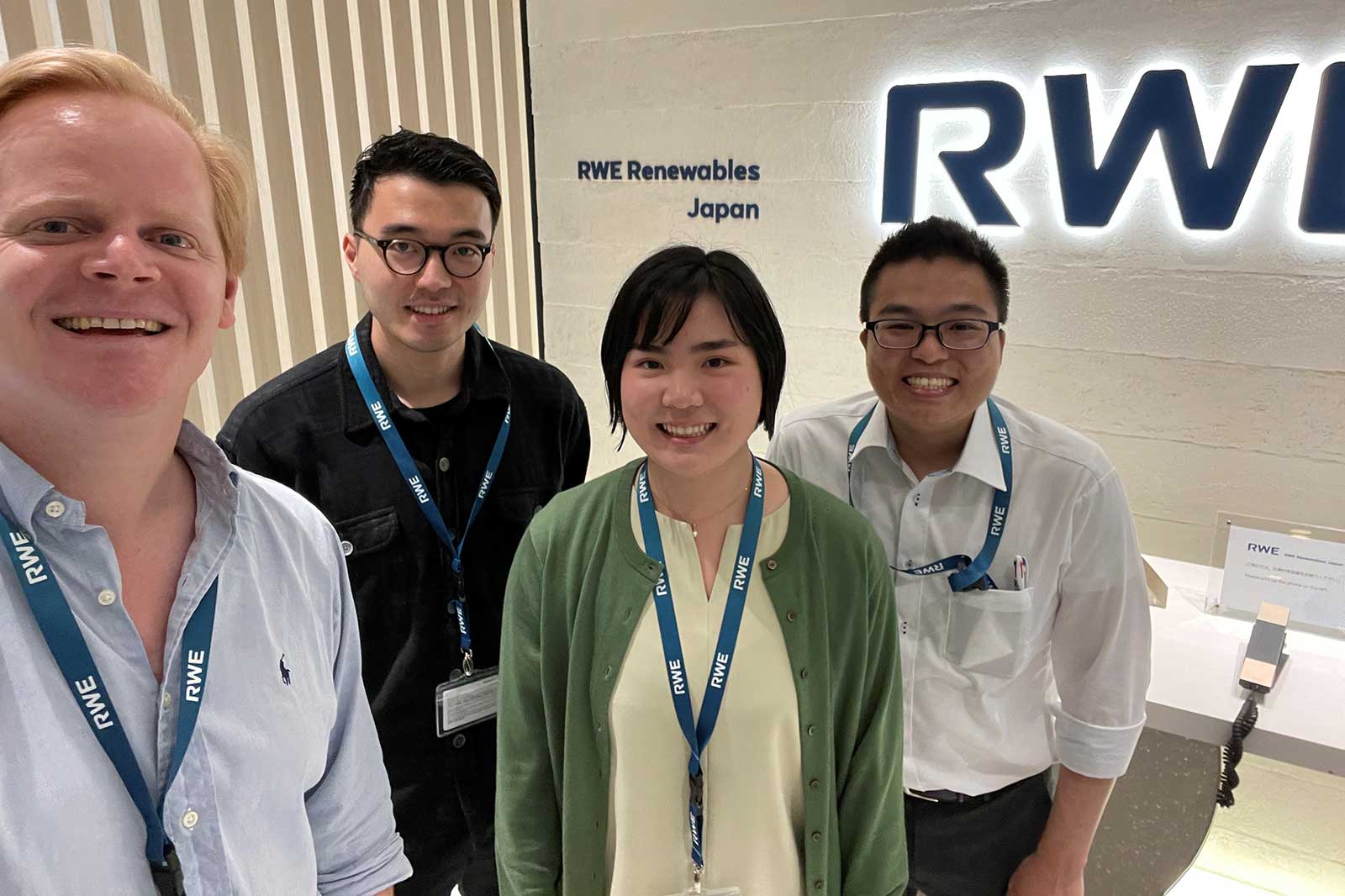 Four individuals stand in front of an RWE Renewables Japan sign, wearing name badges and ID lanyards.