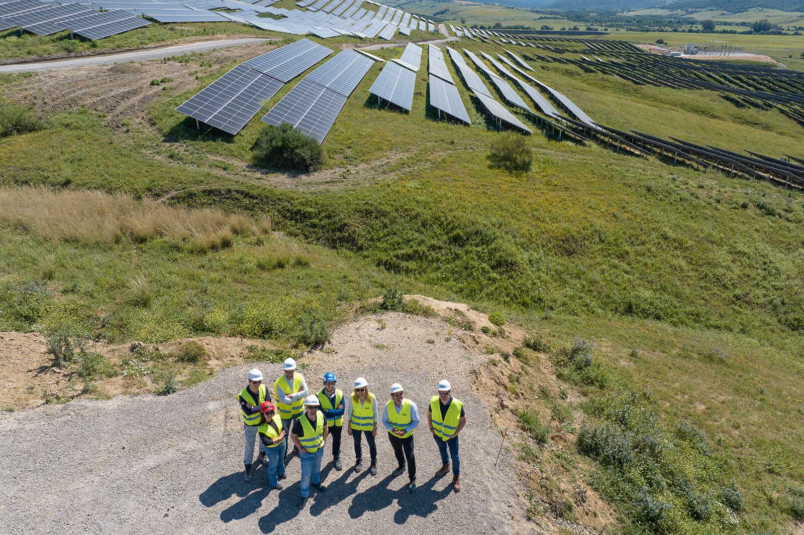 Eine Gruppe von Personen in Warnwesten und Helmen steht auf einem Schotterweg, umgeben von Sonnenkollektoren auf einer grünen Wiese.