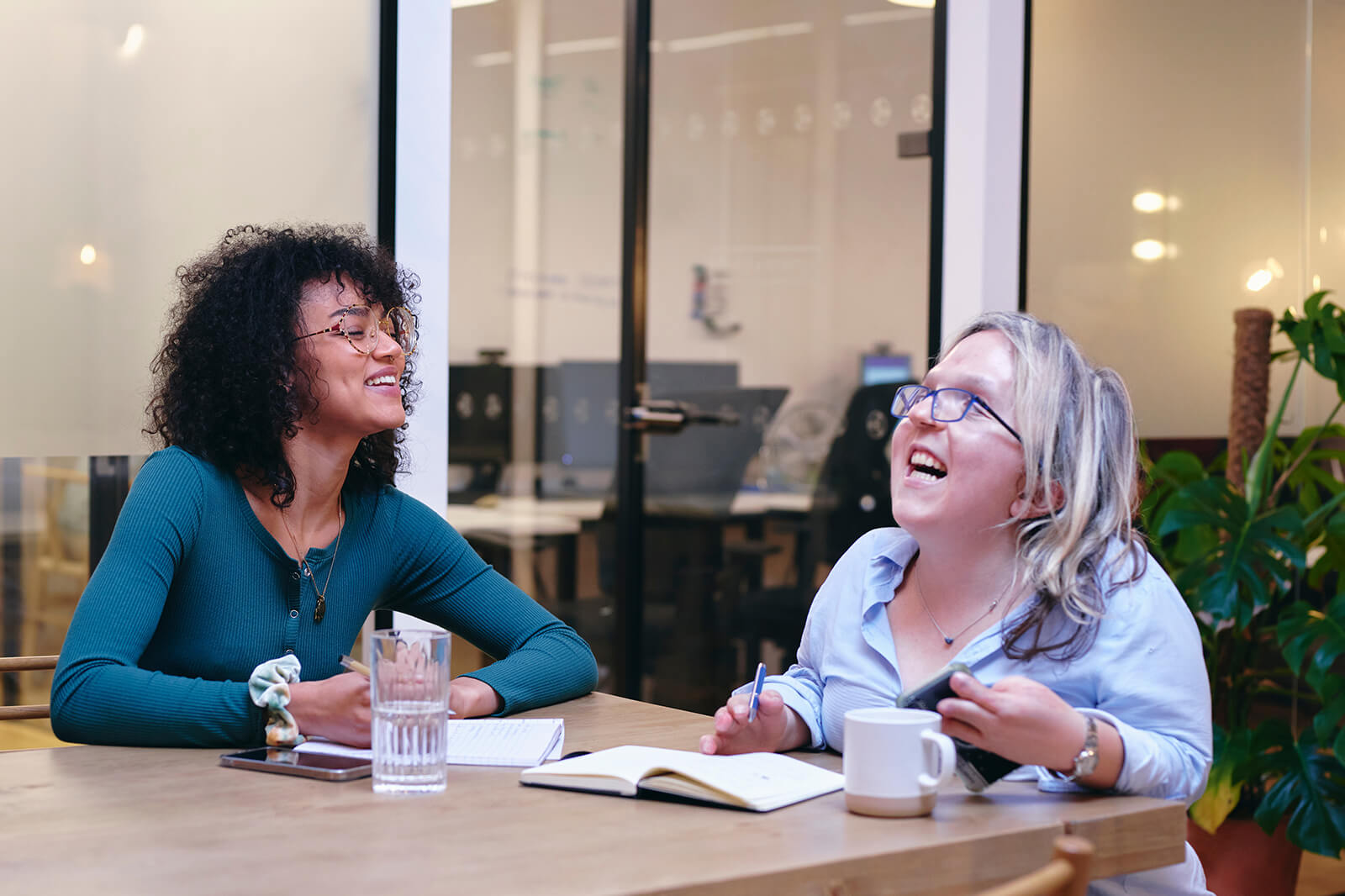 Two women engage in a discussion at a wooden table, with notebooks and a glass of water visible.