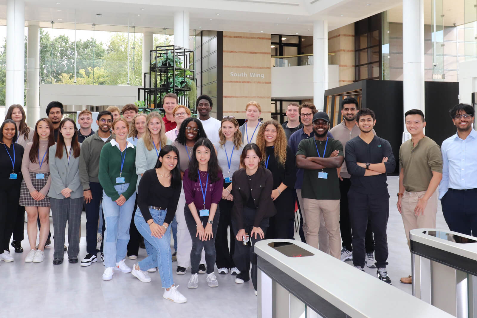 Eine vielfältige Gruppe von Personen steht drinnen zusammen und lächelt für ein Foto in einem modernen Gebäude mit großen Fenstern.